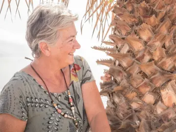 An older couple smiling at each other beside a palm tree by the sea
