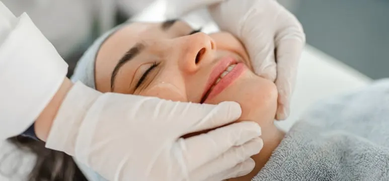 A woman lying back with her eyes closed while a clinician in gloves treats her face