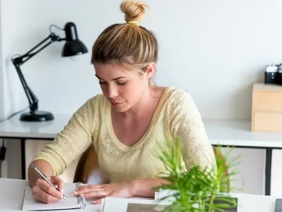 A woman sitting at a desk writing in a notebook beside a lamp