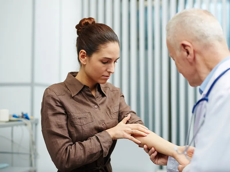 A woman in a brown top showing her forearm to an older doctor in a white coat and stethoscope, in a consulting room