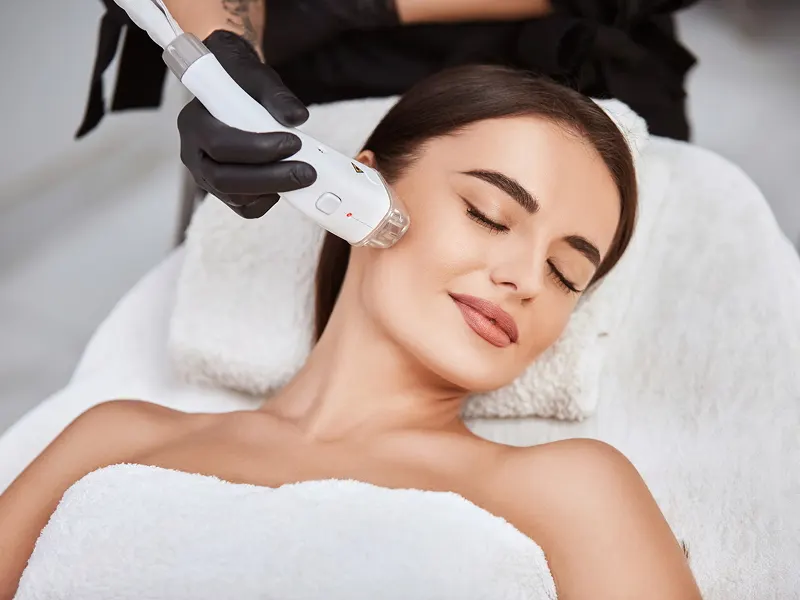 A woman with closed eyes resting on a couch while a gloved hand holds a laser handpiece to her face