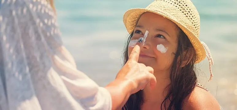 A girl in a straw hat having sunscreen applied to her face by an adult at the beach