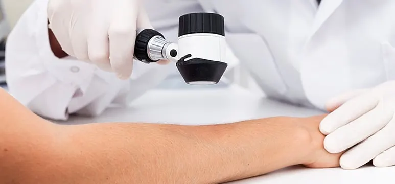 A clinician in a white coat holding a dermatoscope over a patient's forearm