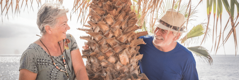 An older couple smiling at each other beside a palm tree by the sea